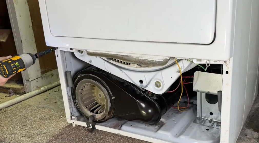 A dryer with its back open, revealing the blower fan, being worked on with a power drill.