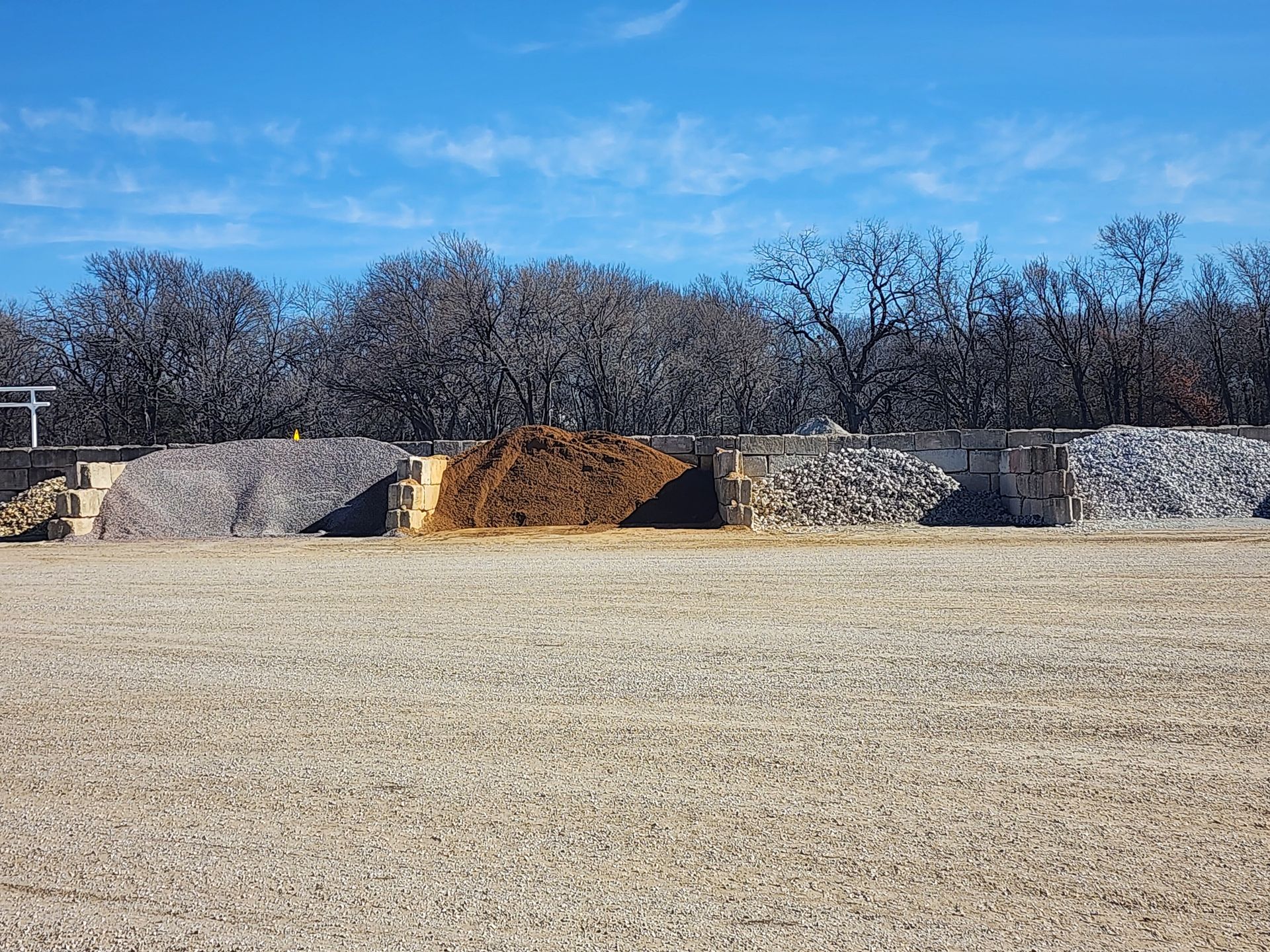 Piles of landscaping materials in an outdoor lot: gravel, mulch, and stone against a backdrop of trees and blue sky.