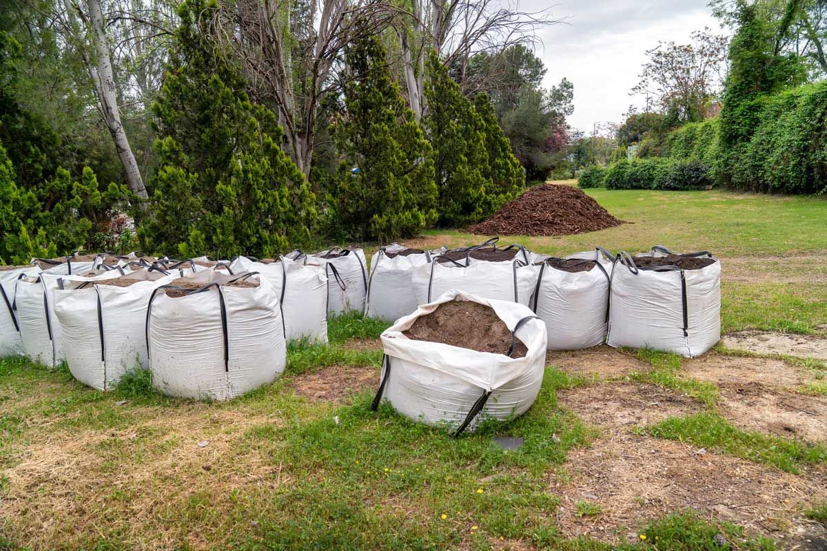 Large white bulk bags filled with topsoil arranged on a grassy property near trees and shrubs.