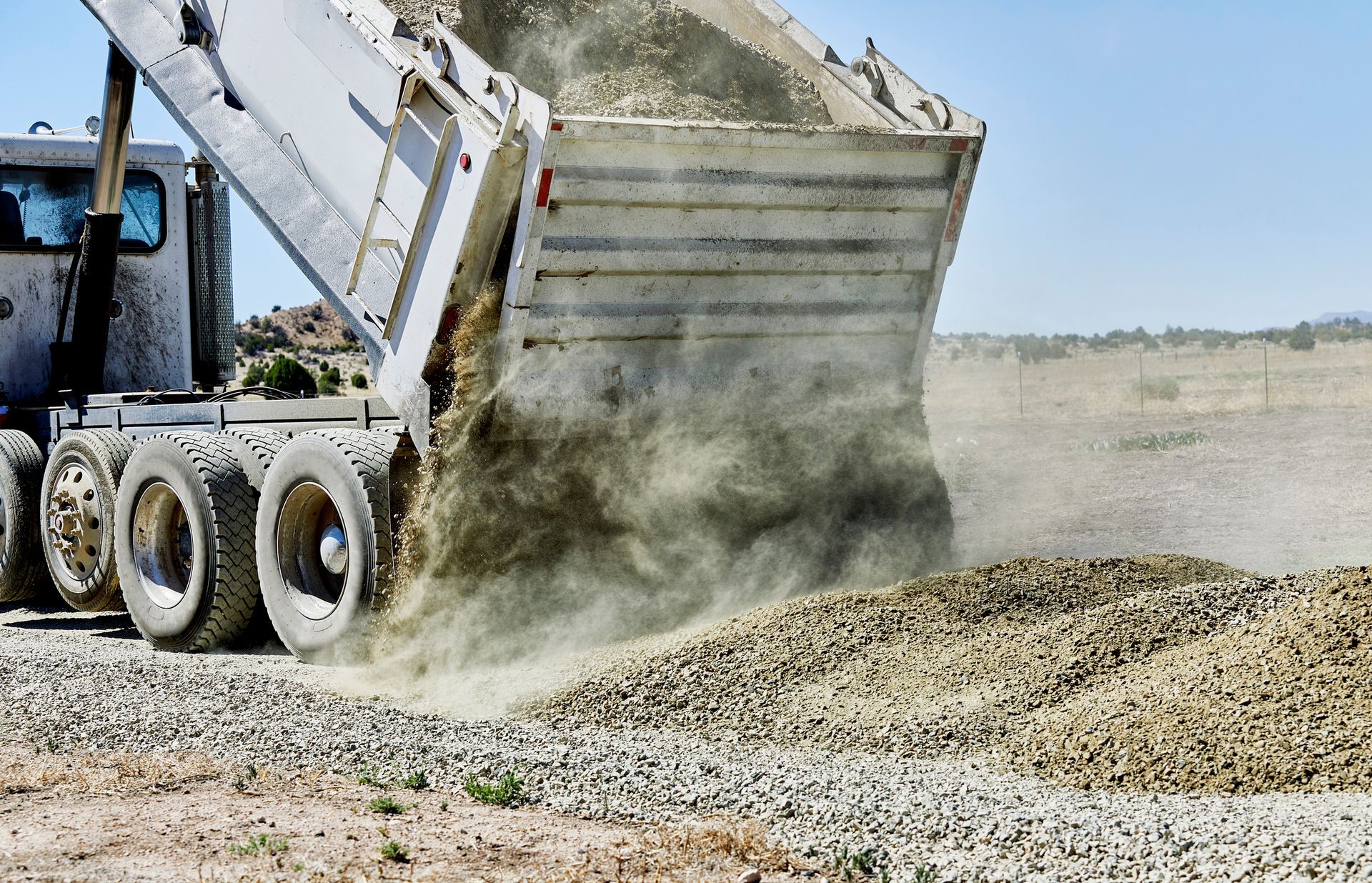 A silver dump truck dumps asphalt on the ground, under the sun.