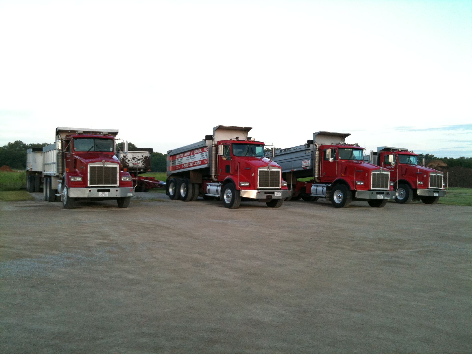 Four red dump trucks parked on a gravel lot; one has a load.