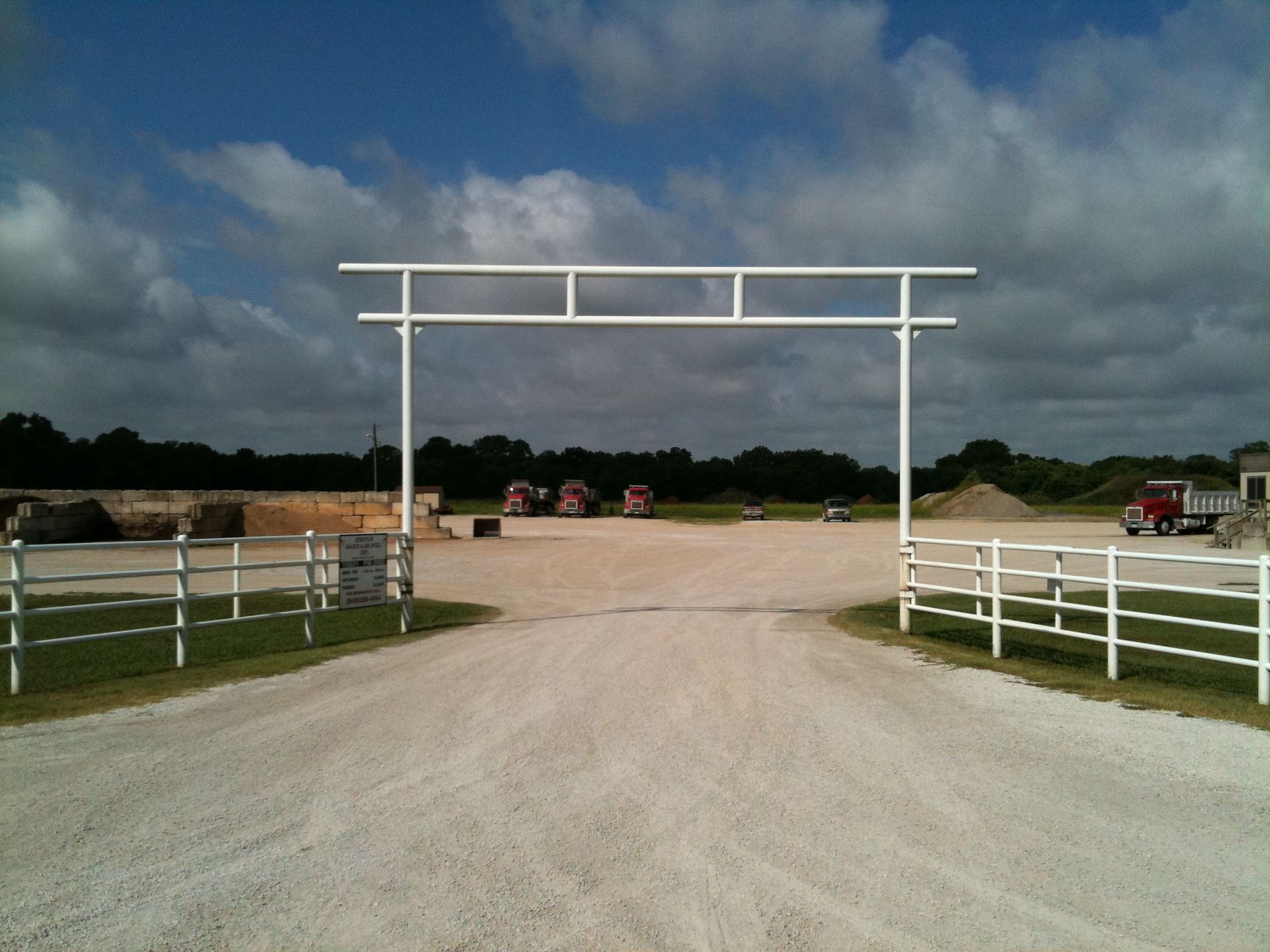 White arched gate over a gravel driveway leading to an industrial site with red trucks.