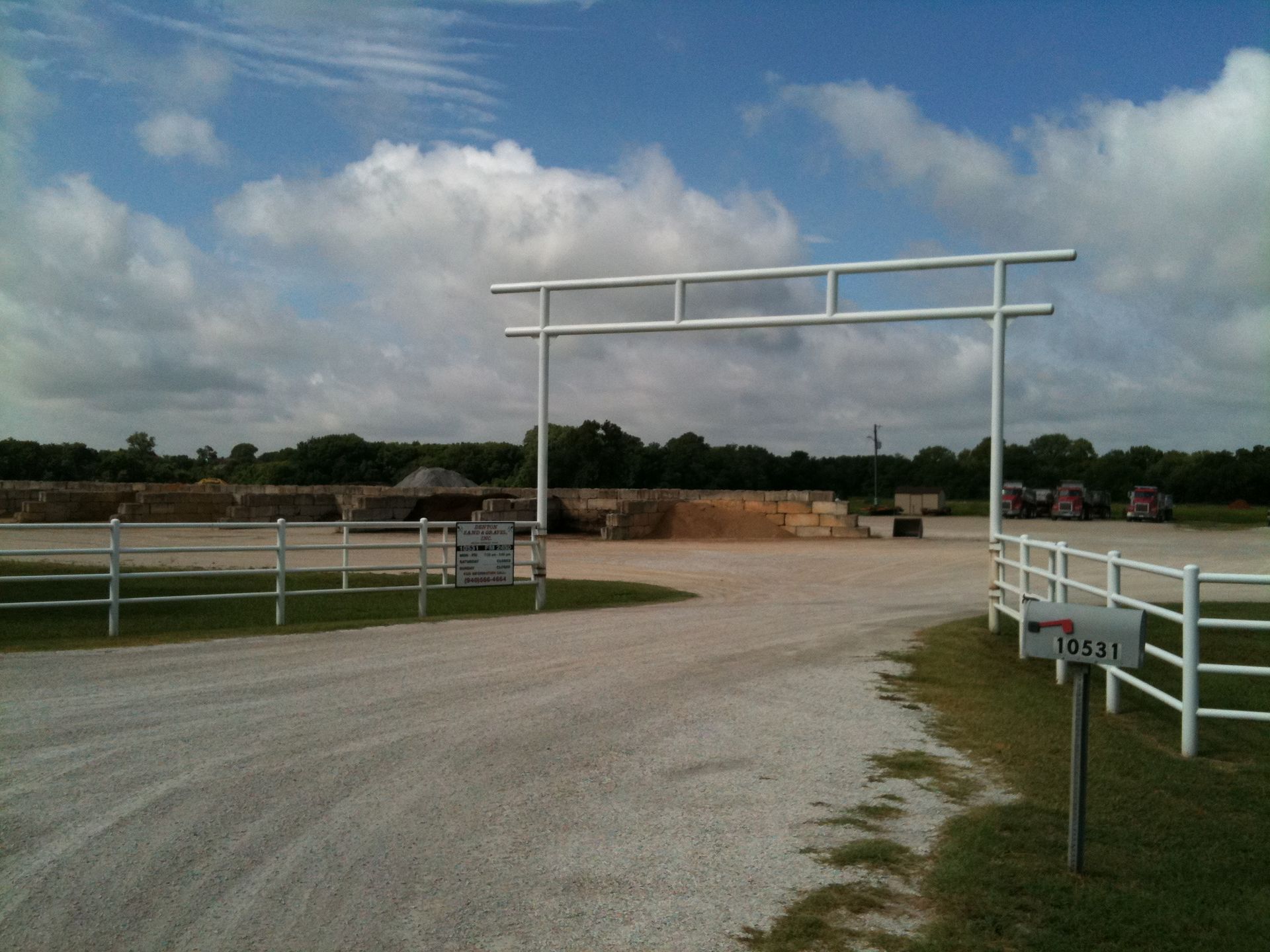 Gravel driveway leads to a white metal gate with stacked materials beyond, under a cloudy sky.