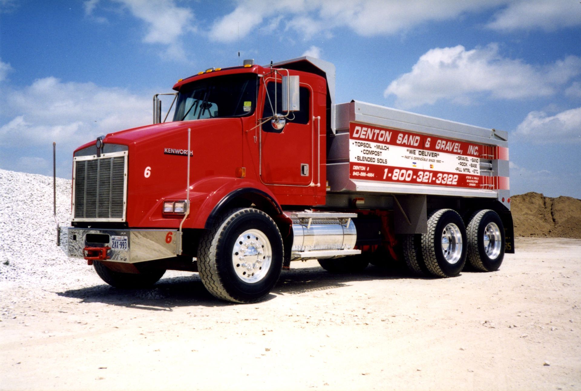Red Kenworth dump truck at Denton Sand & Gravel, on a dirt road under a blue sky.