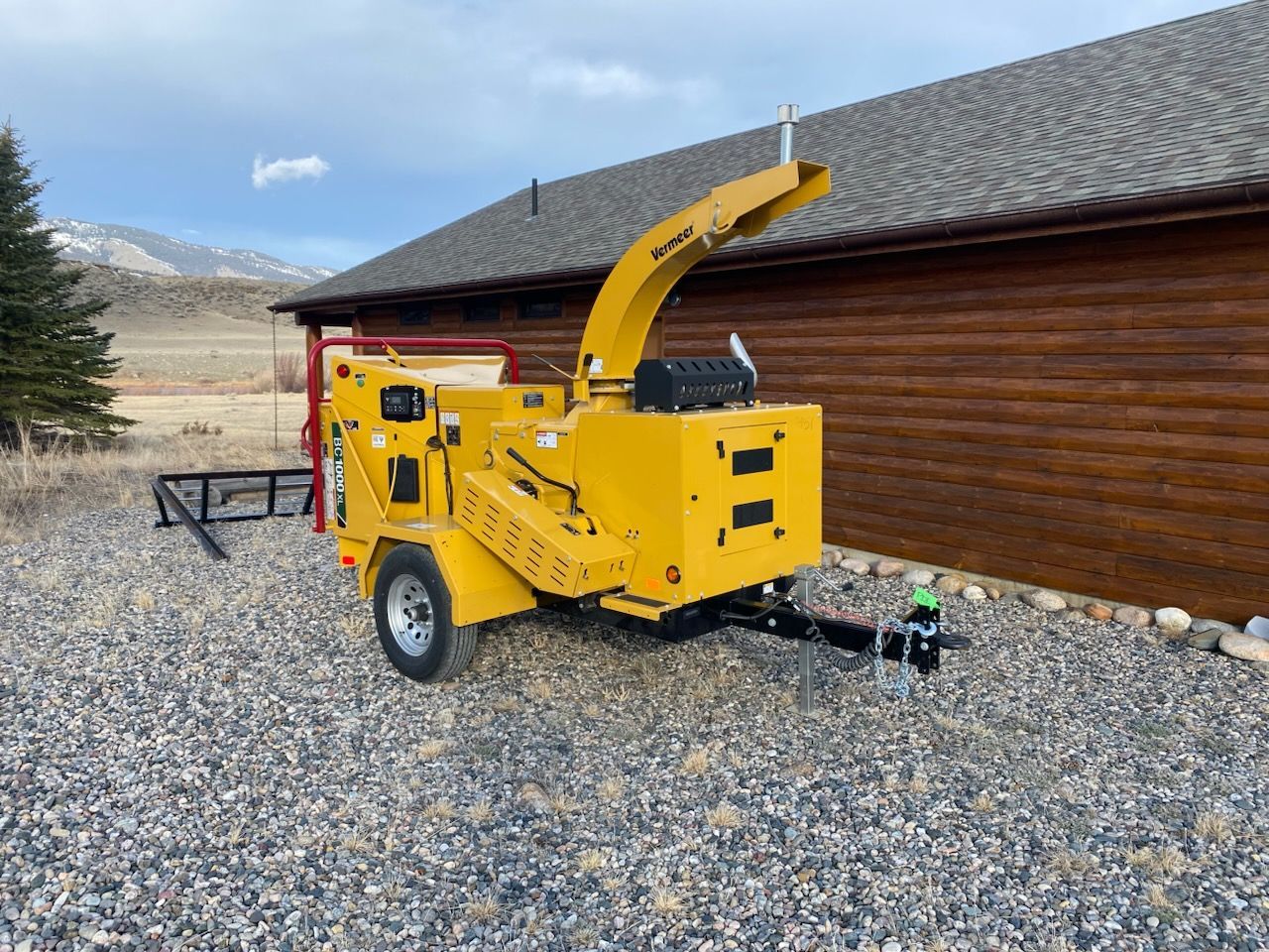 Yellow wood chipper on gravel beside a brown building with snow and mountains in the background