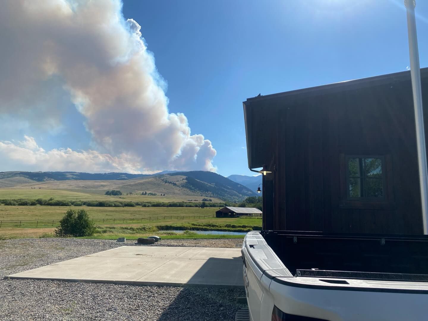 Smoke plume over rural fields beside a dark building under a blue sky