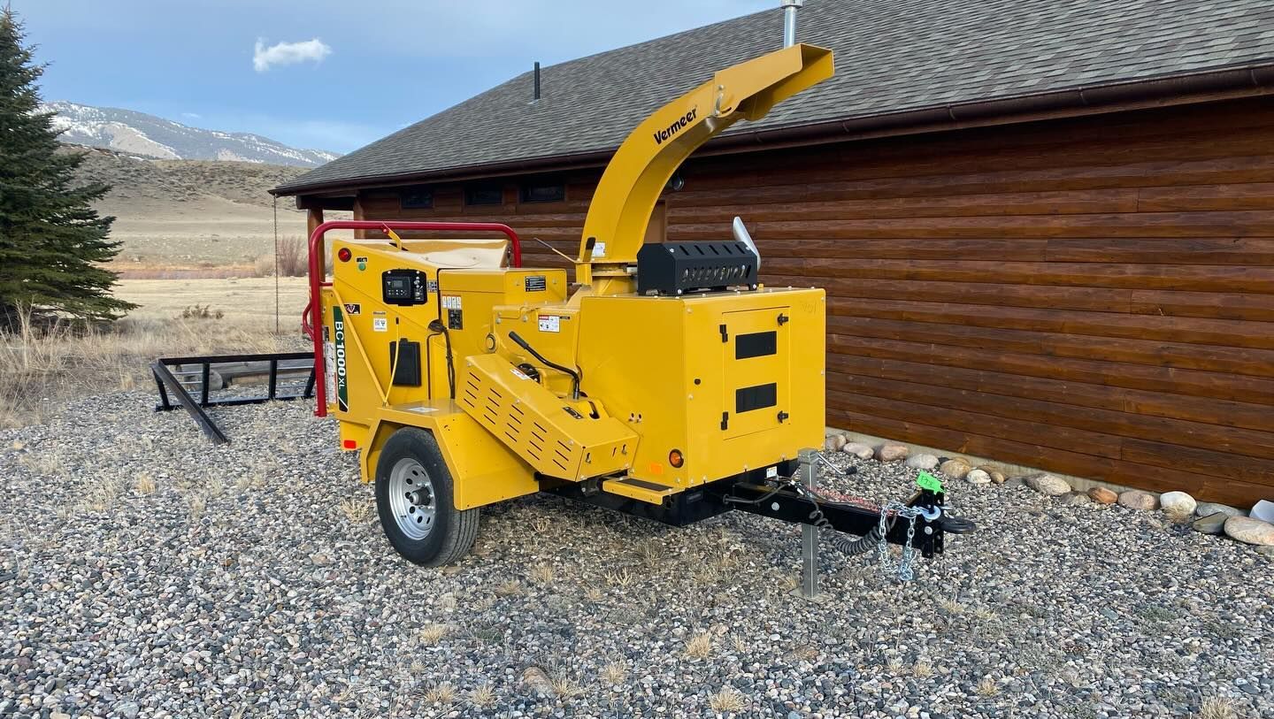 Yellow wood chipper on gravel beside a wooden building, with mountains in the background.