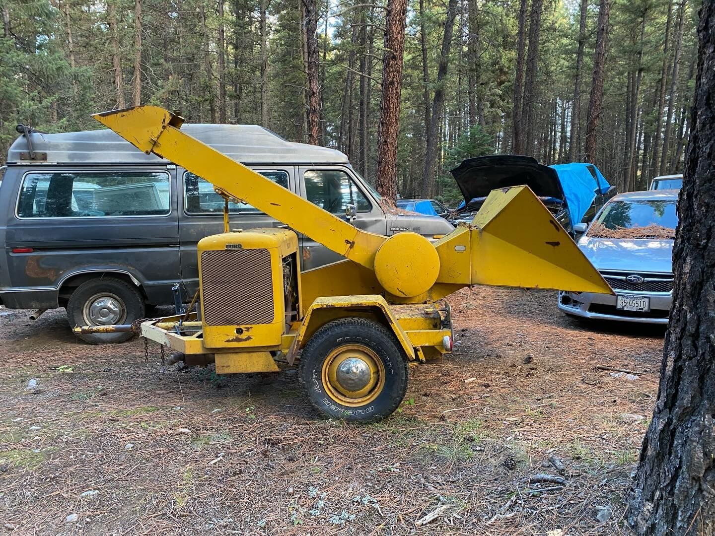 Yellow wood chipper beside parked cars in a forest clearing