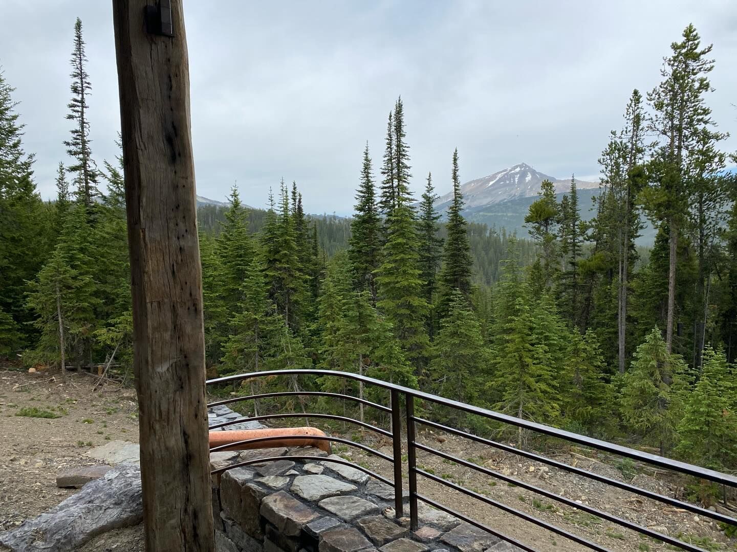 Forest overlook with wooden bench and railing, overlooking evergreen trees and a distant snow-capped mountain.