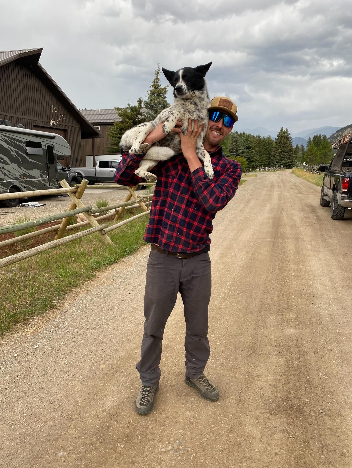 Man holding a black-and-white dog on his shoulders on a gravel road near rustic buildings.