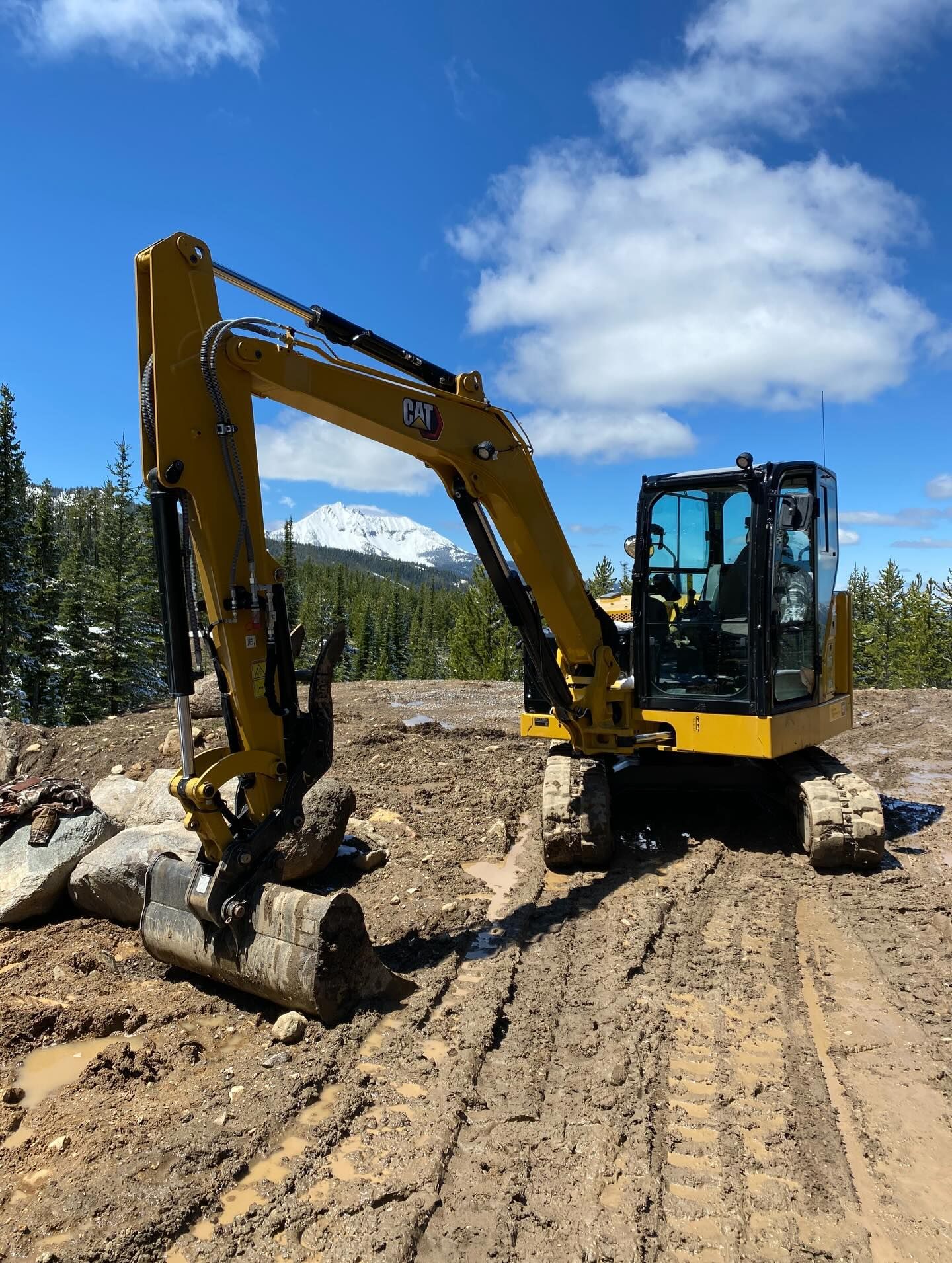 Yellow excavator on a dirt road digging near large rocks under a blue sky.