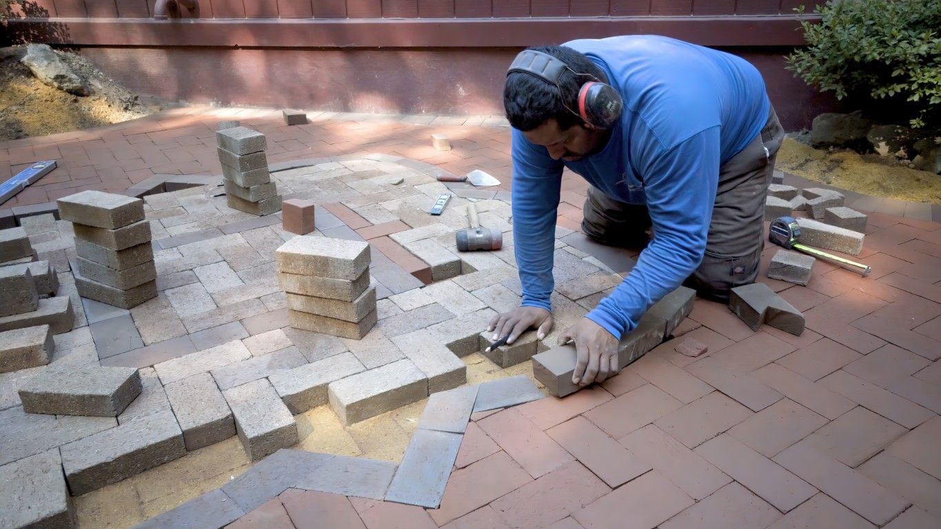 Person laying bricks on a patio, wearing ear protection. Bricks are red and tan. Outdoor setting.