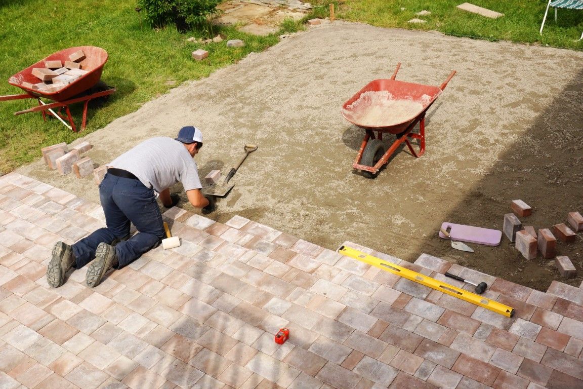 Man laying brick patio, using a level and other tools. Two wheelbarrows sit nearby on gravel base.