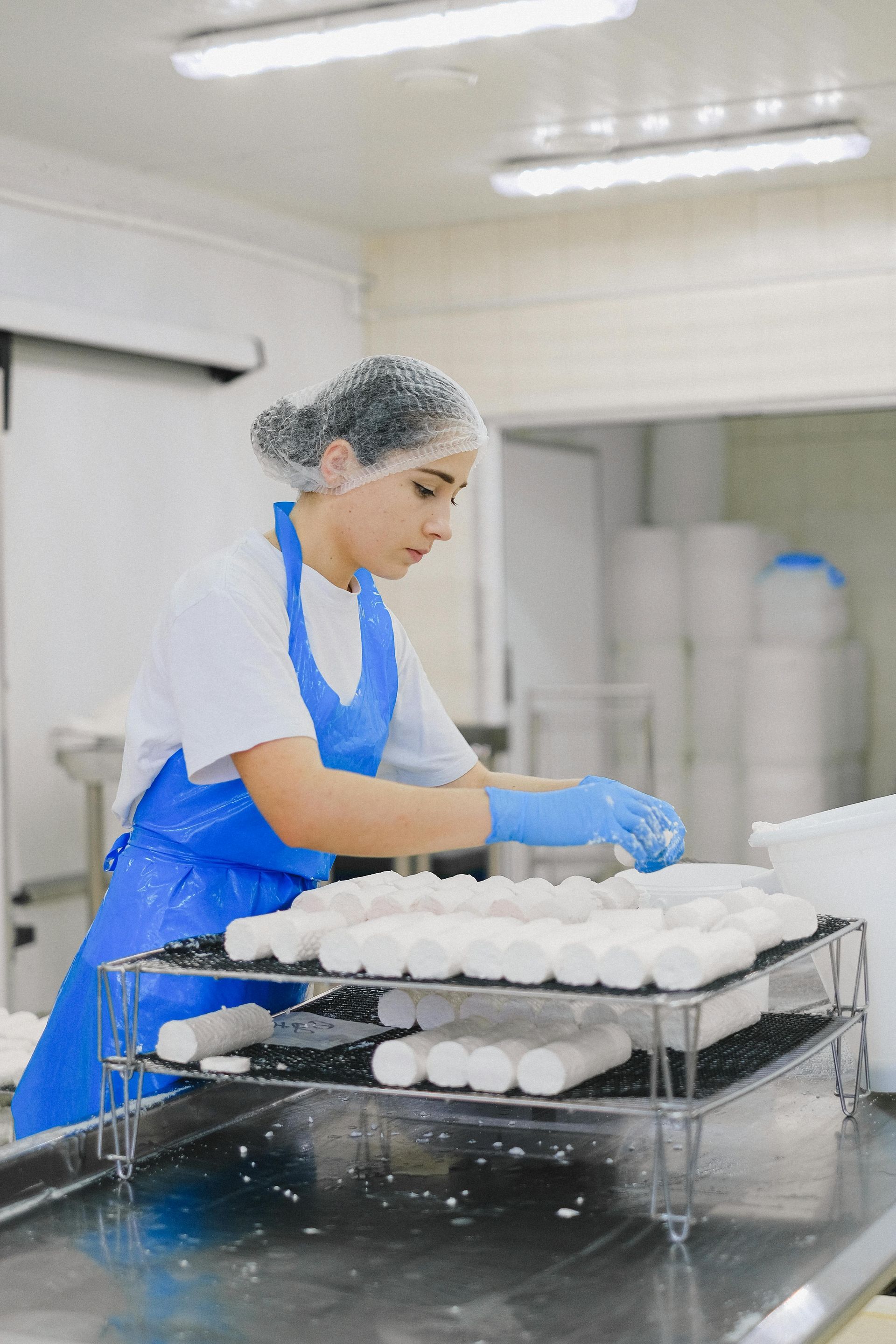 Woman in blue apron and gloves arranging white cheese rolls in a food processing facility.