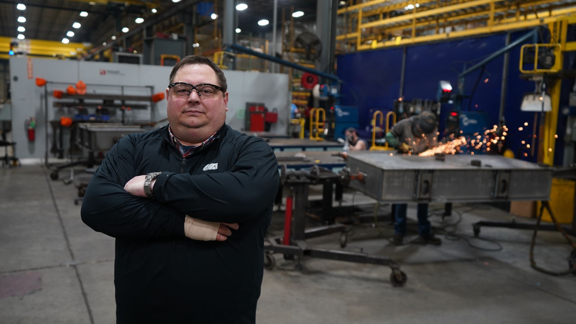 Plant manager posing on the production floor with active metal fabrication and welding operations in the background.