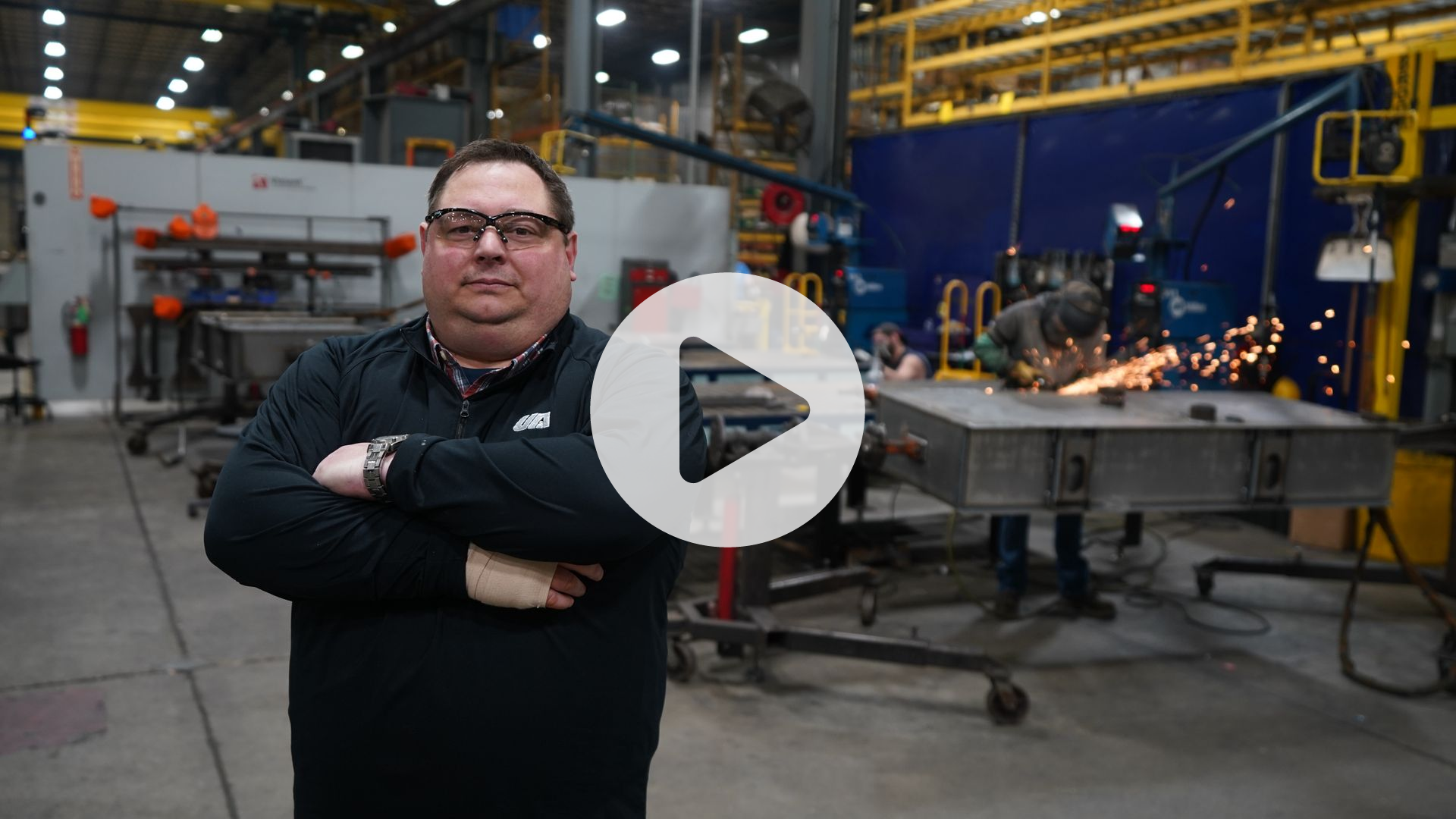 Video thumbnail of a plant manager standing on the factory floor, with workers welding metal in the background.
