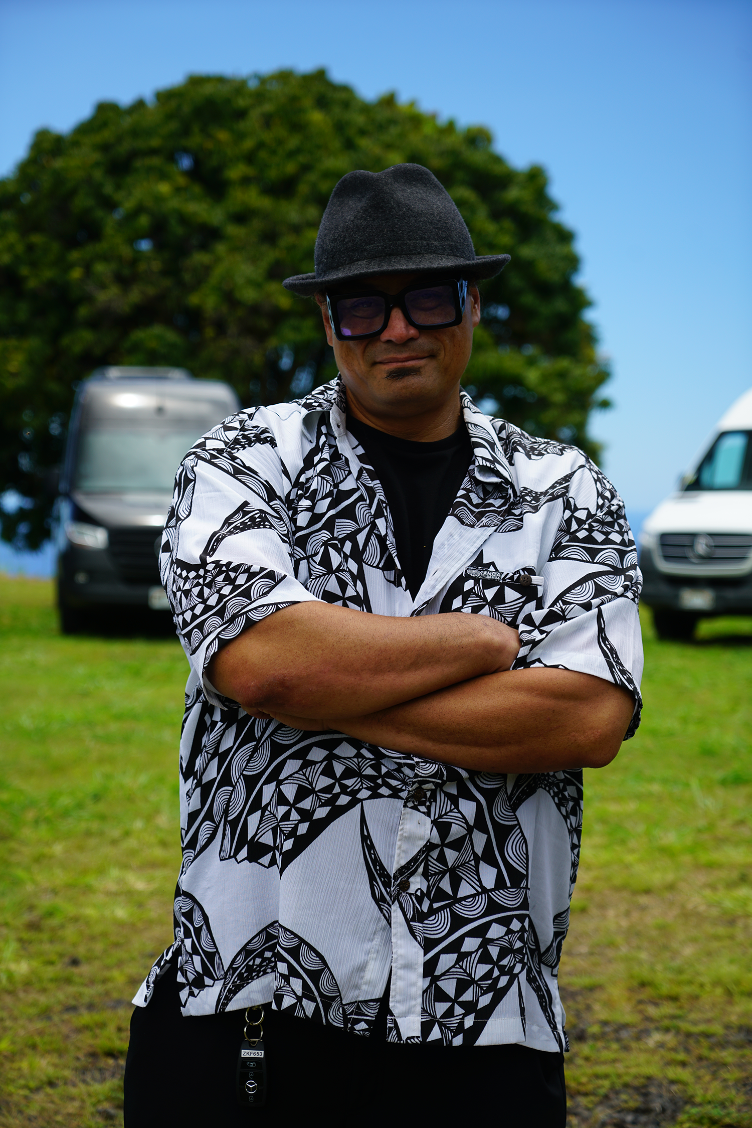 A man wearing a hat and sunglasses is standing in a field with his arms crossed.