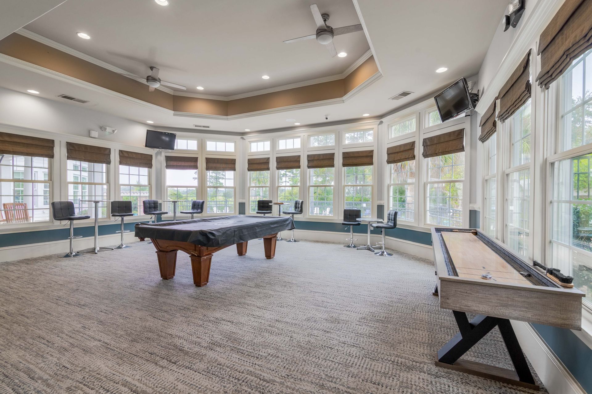 Game room with pool table, shuffleboard, and bar stools. Windows surround the room at Grand Oaks at Ogeechee River in Savannah, GA.