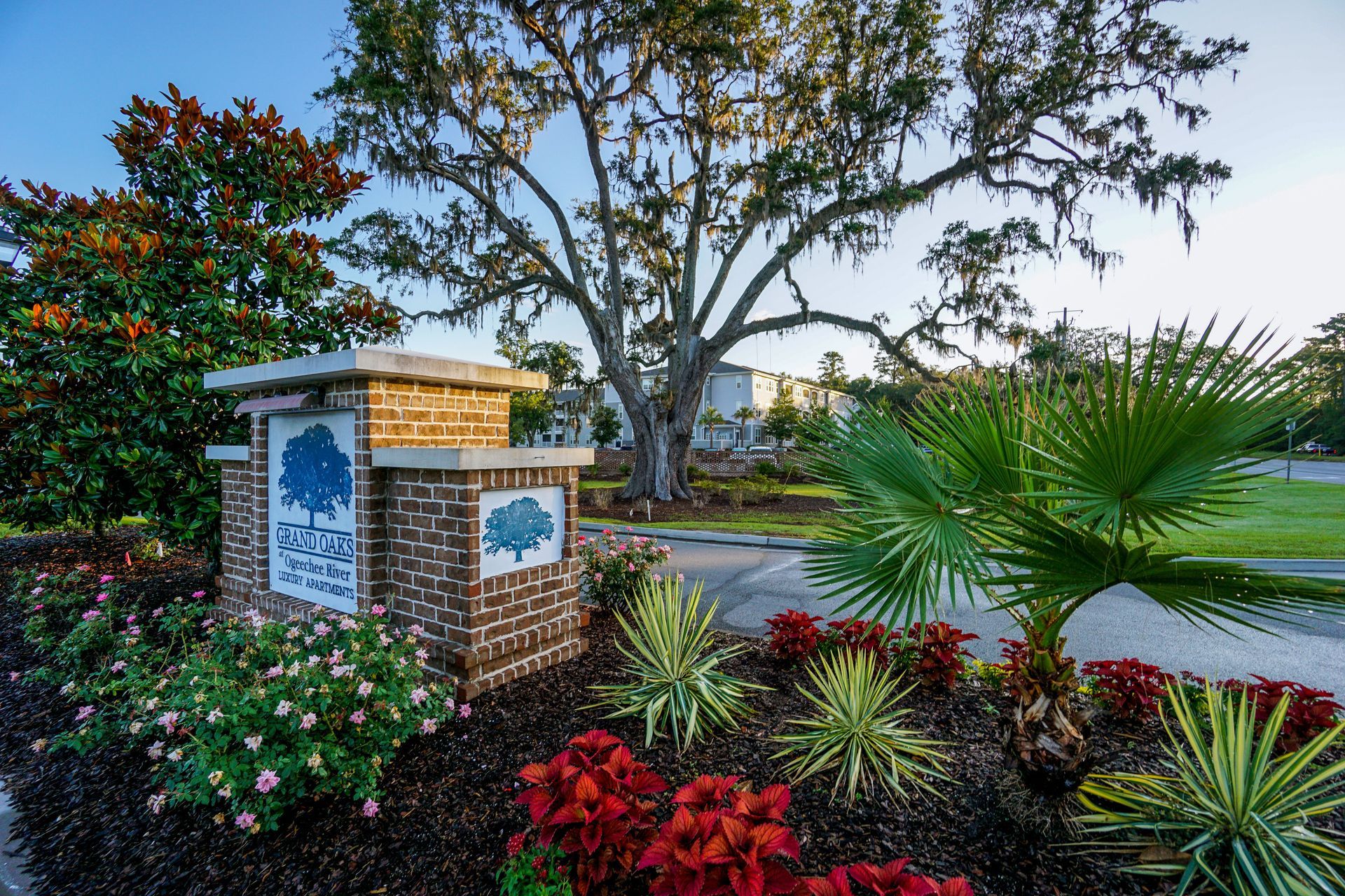 Brick neighborhood entrance sign surrounded by colorful flowers and palm plants beside a street at Grand Oaks at Ogeechee River in Savannah, GA.