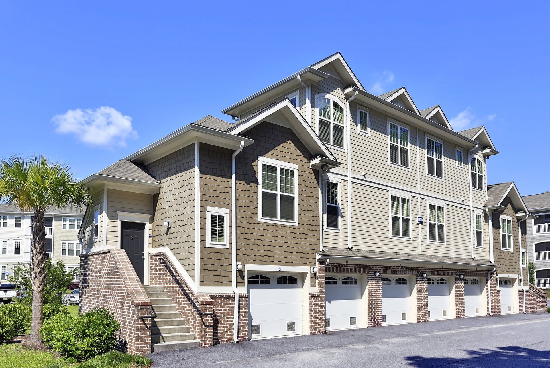 Modern townhouse complex with beige siding, white garages, and stone steps under a blue sky at Grand Oaks at Ogeechee River in GA.