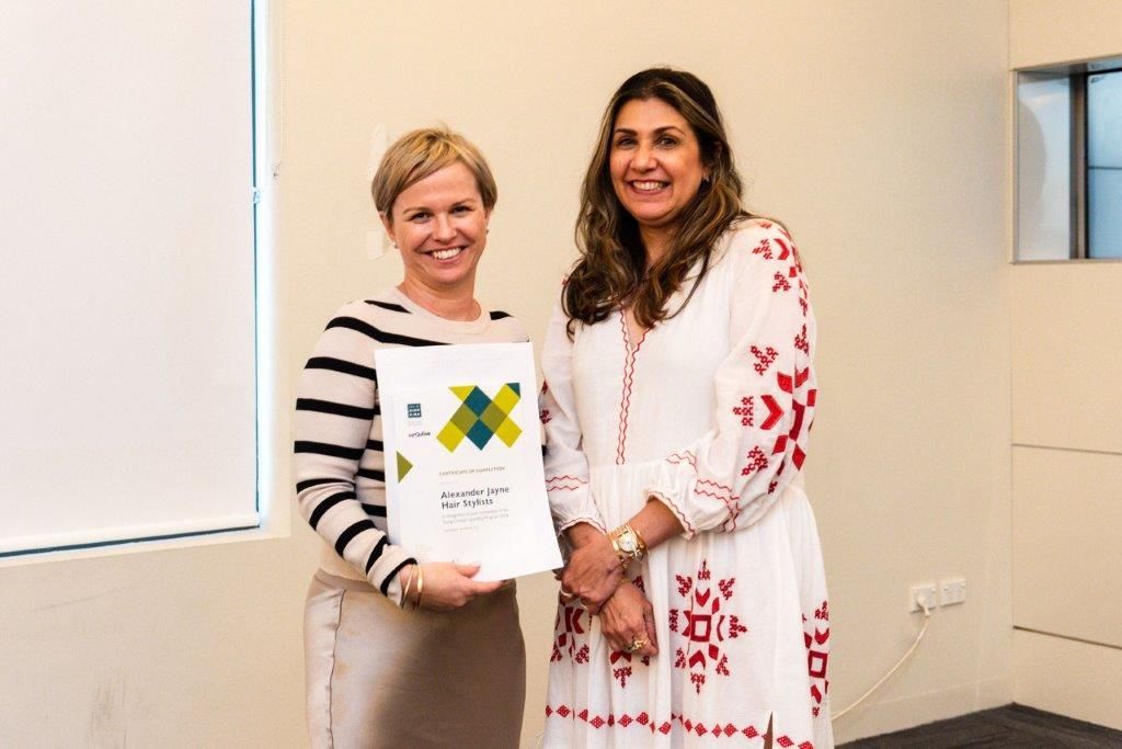 Two women pose, one holds a certificate. Light-colored room, natural light, both smiling.