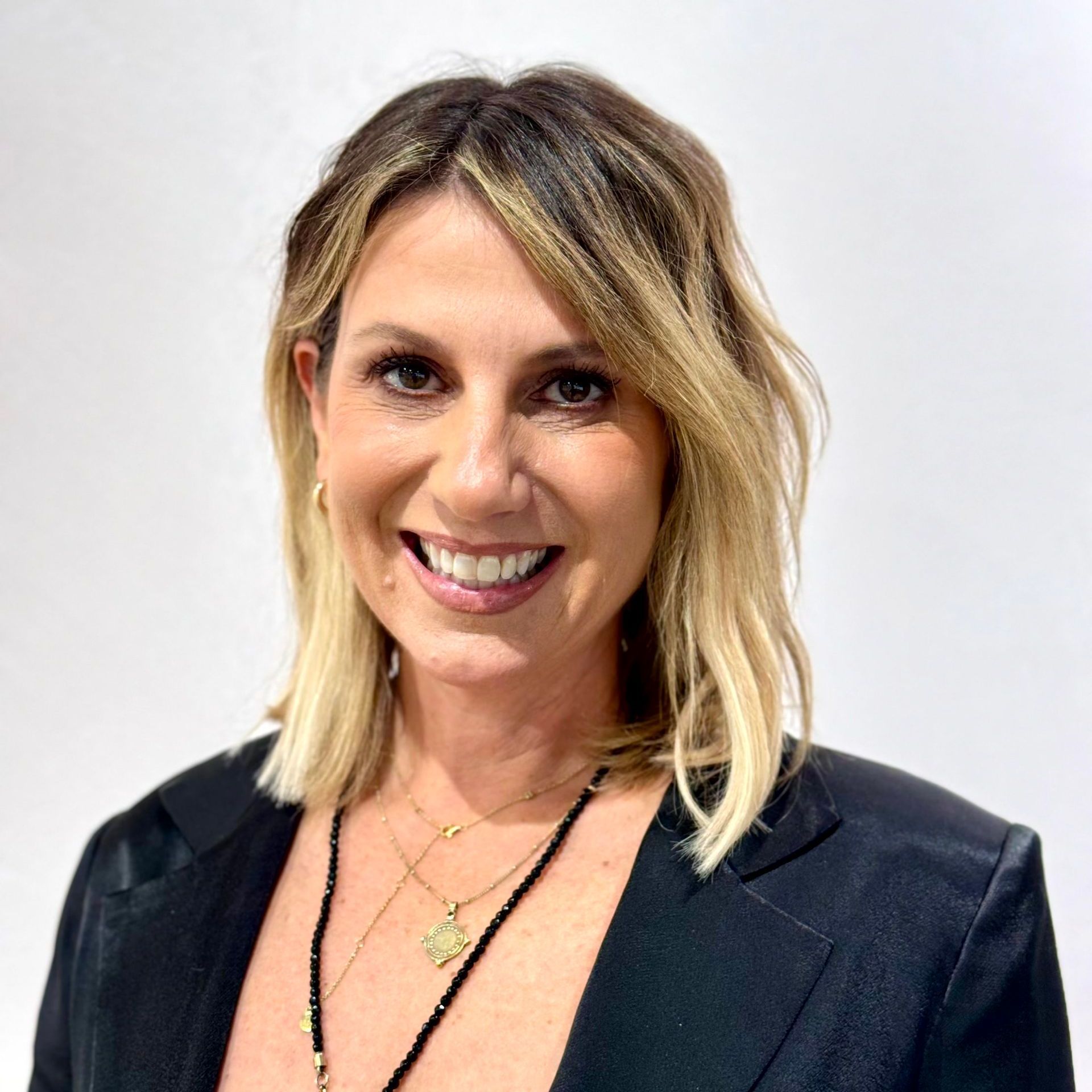 Woman with shoulder-length blonde hair smiling, wearing a black blazer and multiple necklaces in front of a white wall.