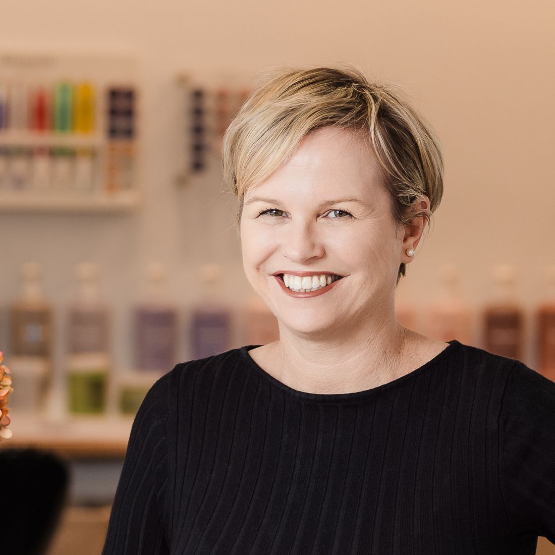 Woman with short blonde hair smiles, wearing a black shirt, in a shop setting with products.
