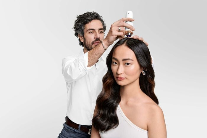 Man uses a device on a woman's scalp. She has long, dark hair and looks at the camera. White backdrop.
