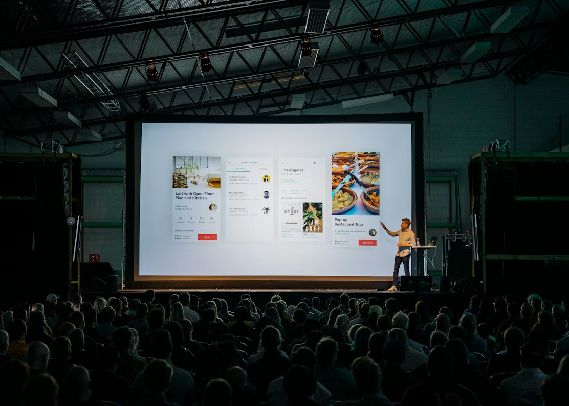 Man presenting app interface on large screen to a seated audience in a large venue.