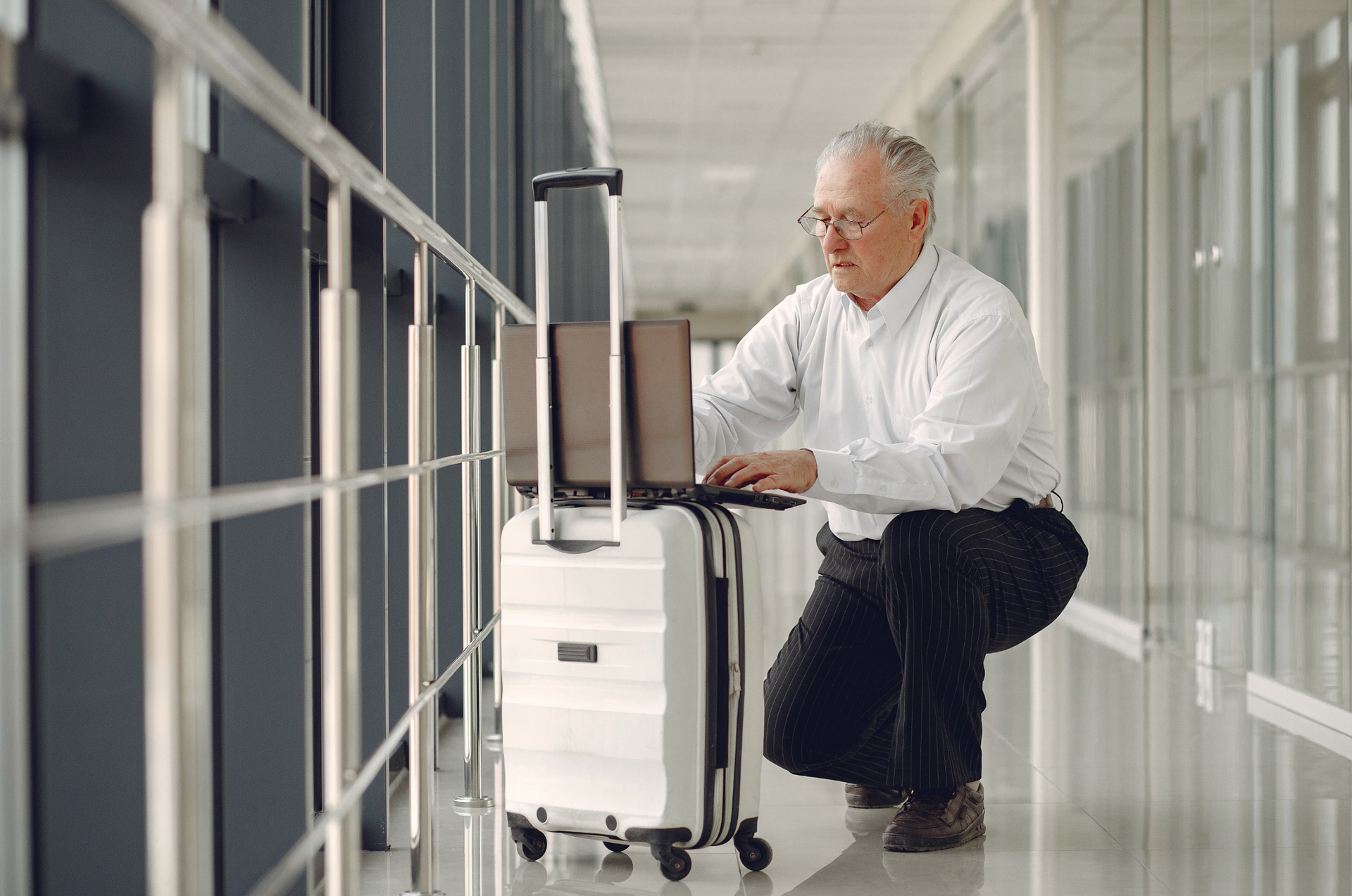 Man crouching beside a white suitcase in a bright hallway, using a laptop on top of it.