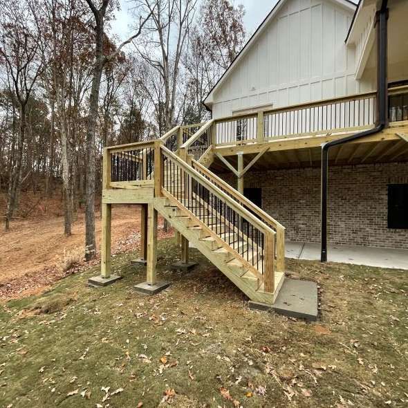 Wooden deck with stairs leading to a white house. Black railings and supports. Outdoors on a cloudy day.