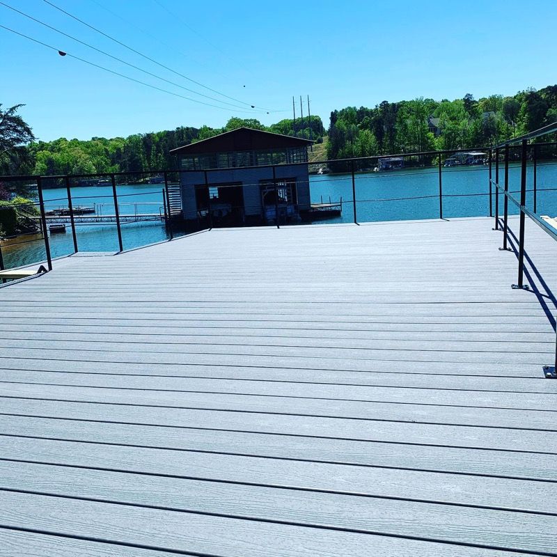 Dock overlooking a lake with a boathouse. Gray deck, blue water, green trees, and clear sky.