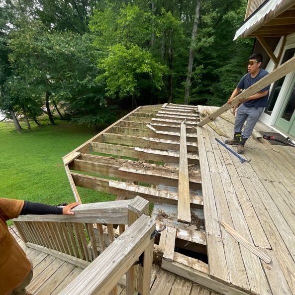 Two people removing decking boards from a partially dismantled wooden deck. Green grass and trees are in the background.