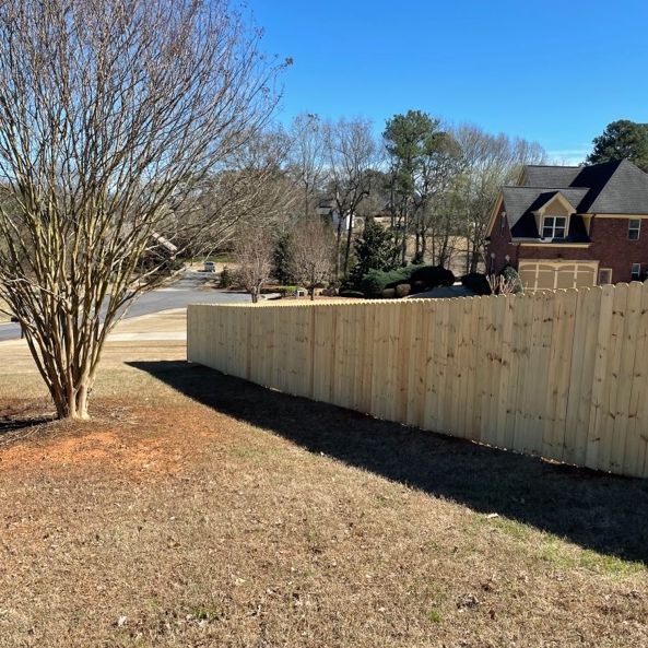 Wooden fence along yard with bare tree, grass, and a house under a clear blue sky.