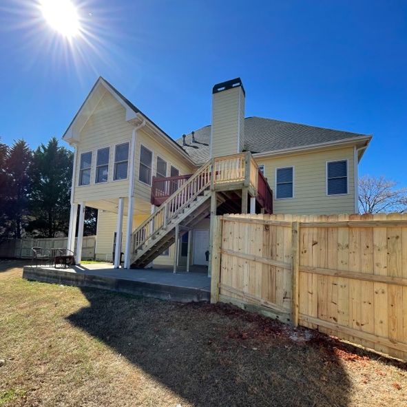 Yellow house with wooden deck and staircase. New wooden fence in front, blue sky overhead.
