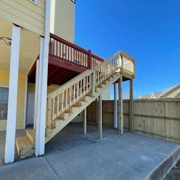 Wooden staircase leading to a red deck on a yellow building. Sunny day, concrete patio below.
