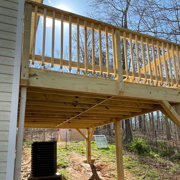 Wooden deck attached to a house, with railing and support beams. Sunlight shines through the trees.