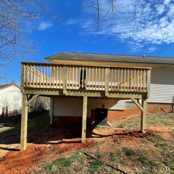 Wooden deck attached to a house with railing, supported by posts. Blue sky, brown earth, and sparse grass.