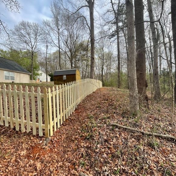 A wooden picket fence curves along the edge of a wooded area, brown leaves cover the ground.