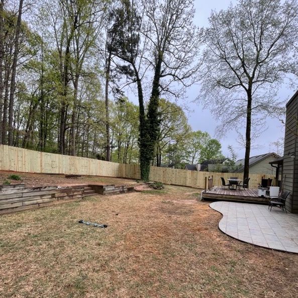 Backyard with newly built wooden fence, trees, deck with furniture, and stone patio.