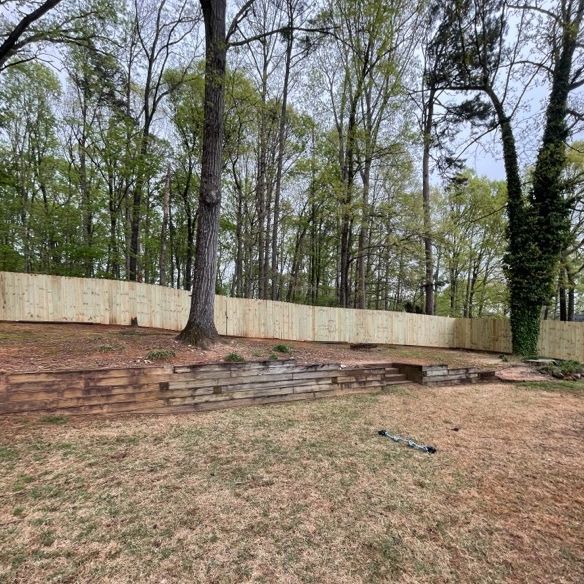 A wooden fence lines a backyard, behind a tiered landscape of wood beams, with trees in the background.