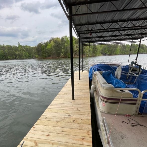 Wooden dock with a pontoon boat covered in blue tarps, under a metal roof, on a lake.