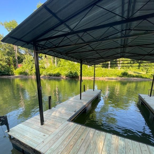 Dock with a black-framed canopy over wooden planks, extending into a body of water with trees in the background.