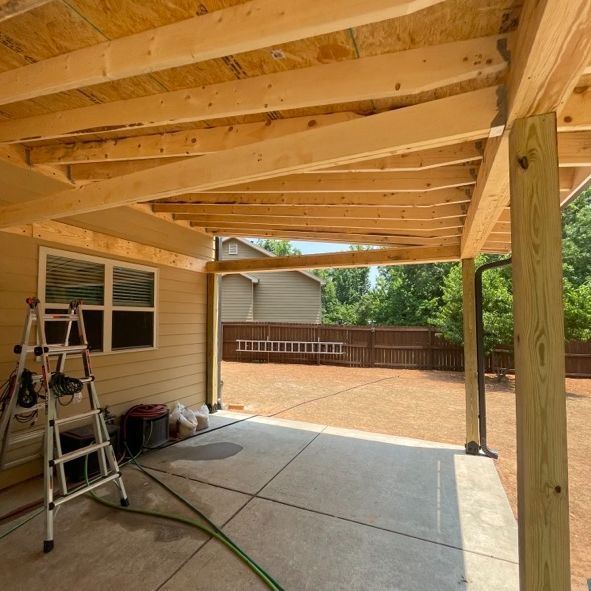 Construction of a covered patio with wooden beams and a concrete floor. A ladder and yard in the background.