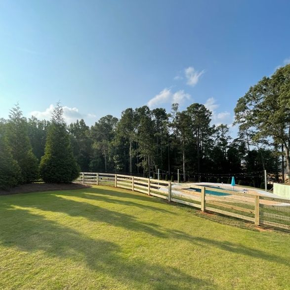 Green lawn with wooden fence and trees under a blue sky, framing a glimpse of a pool.
