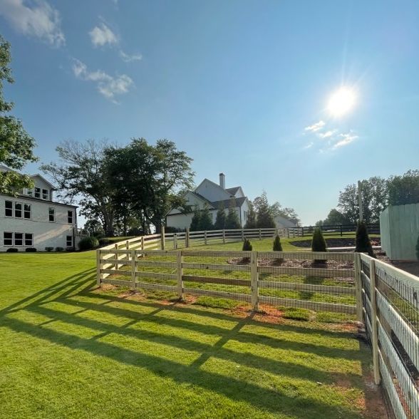 Green grassy yard with white fence and large white house in the background on a sunny day.