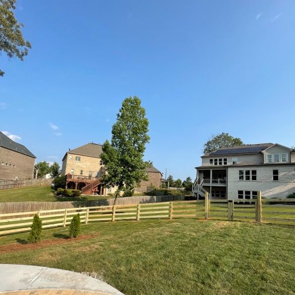 A wooden fence encloses a grassy backyard with a tree. Houses and a bright blue sky are in the background.