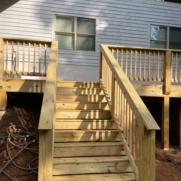 Wooden deck with stairs leading to a house. Railings frame the stairs. Sunlight casts shadows.