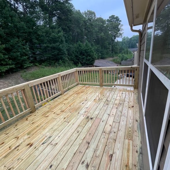 Wooden deck with railing next to a house with large windows. Trees are in the background.