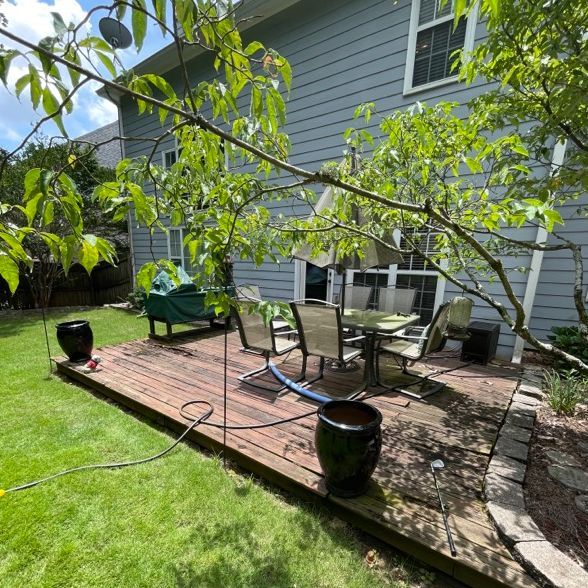 Backyard patio with wooden deck, dining set, and trees against a blue house. Green grass surrounds.